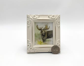 May include: A framed watercolor painting of a moose with large antlers standing in a grassy field. The frame is white with ornate detailing.