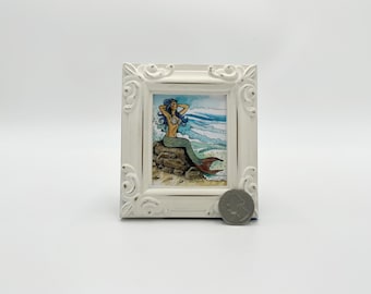 May include: A framed illustration of a mermaid with blue hair and a green tail sitting on a rock. The background is a blue and white watercolor wash depicting the ocean.