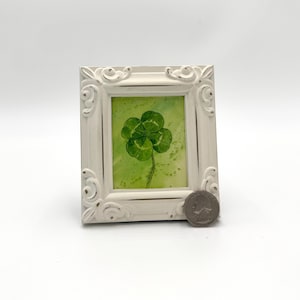 Op de afbeelding: Een wit ingelijst beeld met een groene vierbladige klaver aquarel schilderij erin.