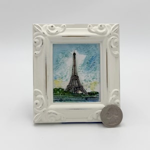 May include: A small, white, ornate picture frame with a painting of the Eiffel Tower. The painting features the tower in brown against a blue and green background. A quarter is next to the frame.