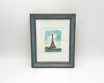 May include: Framed watercolor print of the Eiffel Tower in Paris. The artwork is set within a blue and gold-toned wooden frame. The print depicts the iconic tower against a blue and green background.