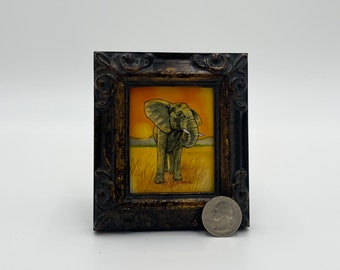 May include: A framed painting of an elephant standing in a field of tall grass. The elephant is facing the left side of the image and has its trunk raised. The background is a sunset with orange and yellow hues. The frame is dark brown with ornate details.
