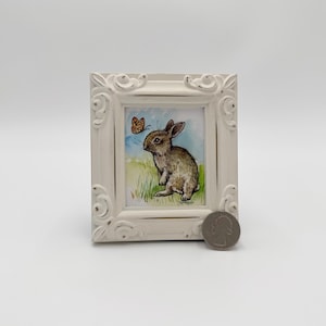 May include: A white framed picture with a brown rabbit looking at a butterfly. The rabbit is in a field of green grass with blue flowers. The butterfly is orange and brown.