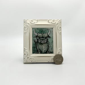 May include: A white framed illustration of a black and white monster with horns and sharp teeth. The monster is set against a green background.