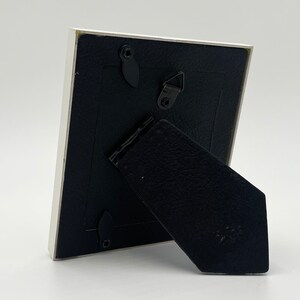 May include: White picture frame with a black backing and a black kickstand. The frame has a small metal hook on the back.
