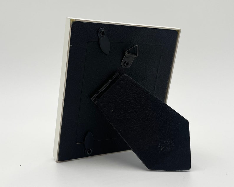 May include: White picture frame with a black backing and a black kickstand. The frame has a small metal hook on the back.