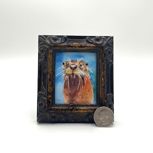 May include: A framed watercolor painting of an otter with a light blue background. The otter is looking directly at the viewer with a curious expression. The frame is dark brown with gold accents.