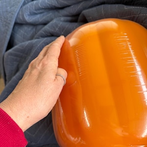May include: A large, smooth, bright orange pumpkin-shaped object with a glossy finish. The surface features vertical indentations that mimic the natural ridges of a pumpkin. The object is rounded and bulbous in form, reflecting light.
