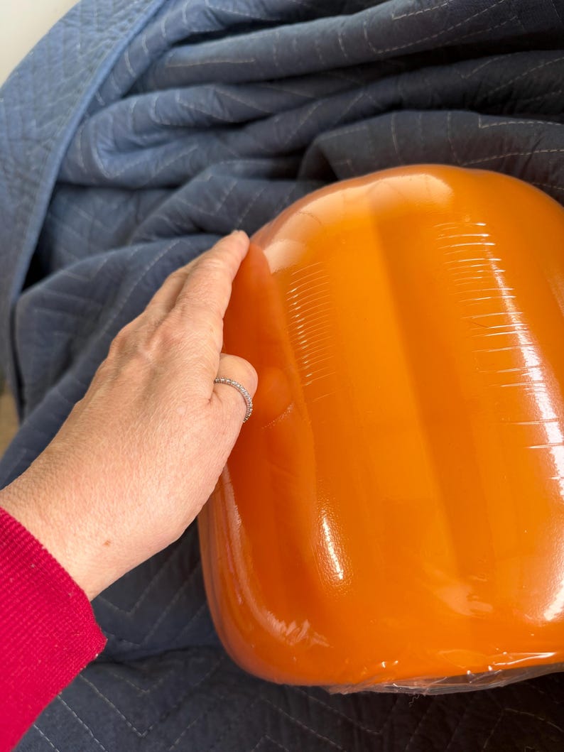 May include: A large, smooth, bright orange pumpkin-shaped object with a glossy finish. The surface features vertical indentations that mimic the natural ridges of a pumpkin. The object is rounded and bulbous in form, reflecting light.