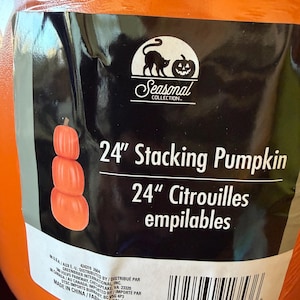 May include: A bright orange 24-inch stacking pumpkin decoration wrapped in clear plastic. The black product label features a cat and jack-o'-lantern logo, the text 24" Stacking Pumpkin, 24" Citrouilles empilables, and a barcode at the bottom.
