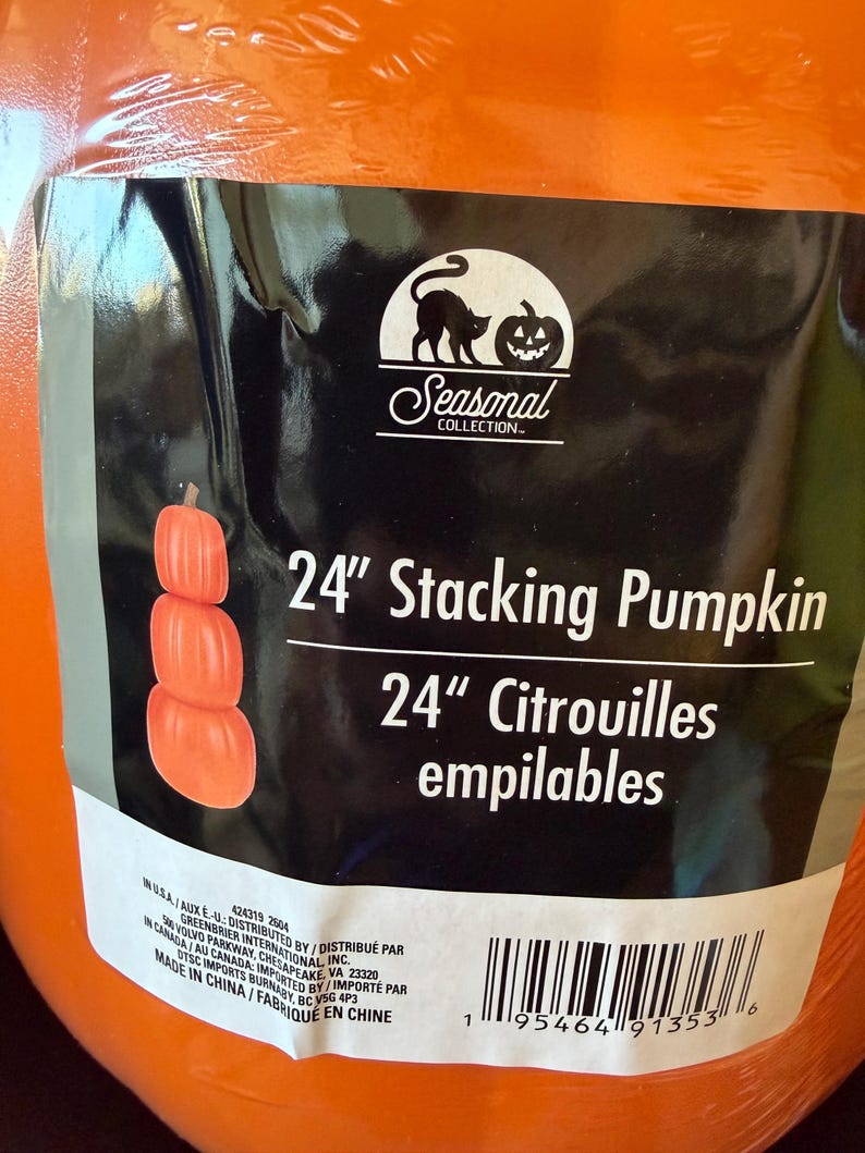 May include: A bright orange 24-inch stacking pumpkin decoration wrapped in clear plastic. The black product label features a cat and jack-o'-lantern logo, the text 24" Stacking Pumpkin, 24" Citrouilles empilables, and a barcode at the bottom.