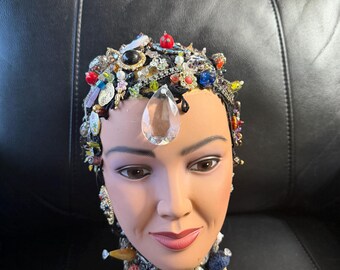 May include: A mannequin head adorned with a jeweled crown and necklace. The crown features an array of colorful gems and beads, with a large clear teardrop-shaped stone at the forehead. The necklace is a wide, ornate band with various jewels.
