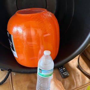May include: A bright orange, spherical plastic container wrapped in clear protective film, positioned inside a large black basin. A clear plastic bottle of purified water is placed in the foreground for scale, next to a small black remote control.