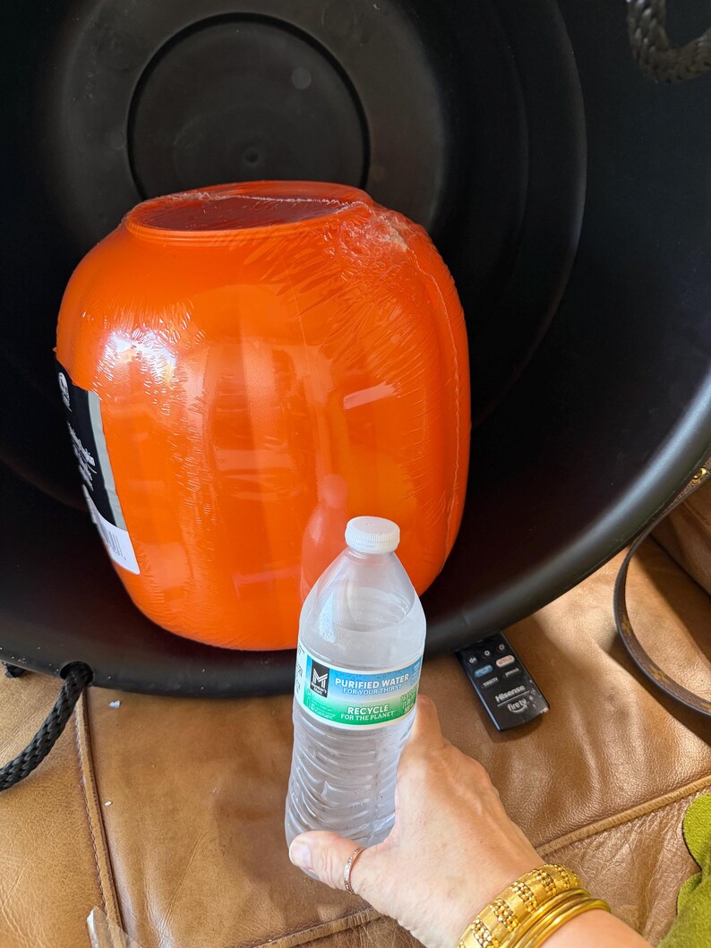 May include: A bright orange, spherical plastic container wrapped in clear protective film, positioned inside a large black basin. A clear plastic bottle of purified water is placed in the foreground for scale, next to a small black remote control.
