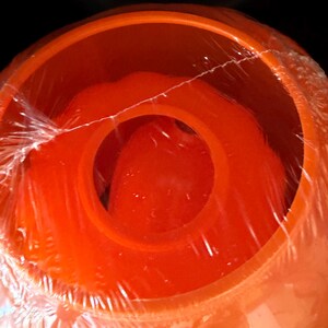 May include: A bright orange, circular plastic container with a wide, open top and a central circular opening. The object is wrapped in clear, thin plastic film, which creates visible wrinkles and reflections across the smooth orange surface.