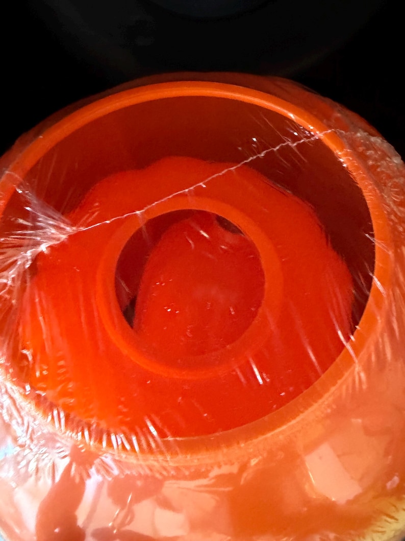 May include: A bright orange, circular plastic container with a wide, open top and a central circular opening. The object is wrapped in clear, thin plastic film, which creates visible wrinkles and reflections across the smooth orange surface.
