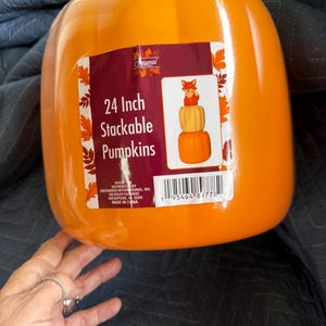 May include: A bright orange plastic pumpkin container featuring a rectangular product label. The label displays the text 24 Inch Stackable Pumpkins and an illustration of stacked pumpkins topped with a fox. A barcode is printed at the bottom.