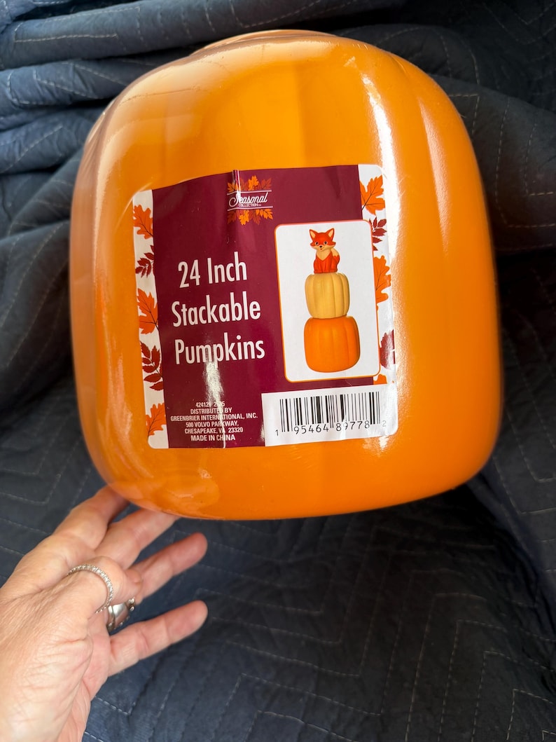 May include: A bright orange plastic pumpkin container featuring a rectangular product label. The label displays the text 24 Inch Stackable Pumpkins and an illustration of stacked pumpkins topped with a fox. A barcode is printed at the bottom.