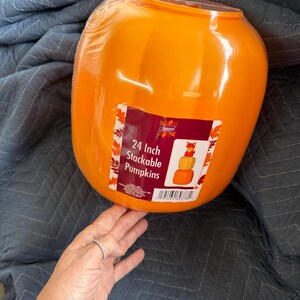 May include: A bright orange plastic pumpkin container with a glossy finish. A rectangular product label is attached to the front, featuring the text 24 Inch Stackable Pumpkins and an illustration of stacked pumpkins topped with a small fox figure.