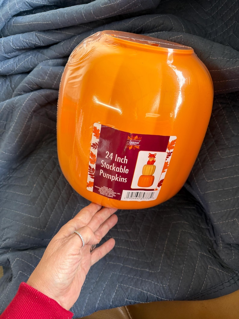 May include: A bright orange plastic pumpkin container with a glossy finish. A rectangular product label is attached to the front, featuring the text 24 Inch Stackable Pumpkins and an illustration of stacked pumpkins topped with a small fox figure.
