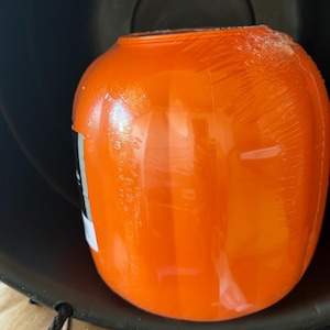 May include: A bright orange, rounded plastic container with a smooth, glossy surface, partially wrapped in clear protective plastic film. The container features a wide opening at the top and a black label on the side with partially visible white text.