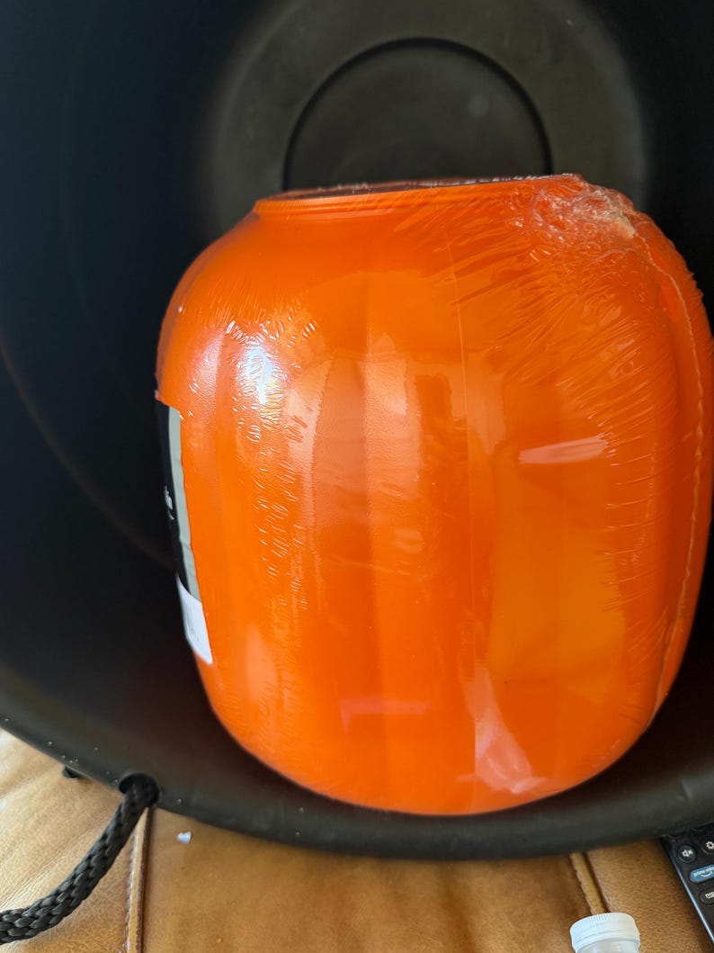 May include: A bright orange, rounded plastic container with a smooth, glossy surface, partially wrapped in clear protective plastic film. The container features a wide opening at the top and a black label on the side with partially visible white text.
