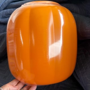 May include: A glossy, bright orange glass pumpkin with vertical ribbed detailing. The surface is smooth and reflective, showcasing a rounded, bulbous shape that mimics the natural form of a pumpkin. The glass has a vibrant, solid orange hue.
