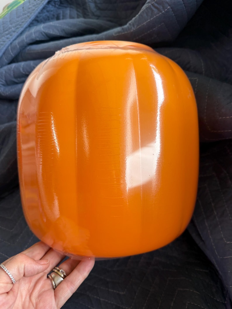 May include: A glossy, bright orange glass pumpkin with vertical ribbed detailing. The surface is smooth and reflective, showcasing a rounded, bulbous shape that mimics the natural form of a pumpkin. The glass has a vibrant, solid orange hue.