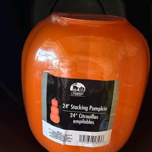 May include: A bright orange, rounded plastic pumpkin decoration wrapped in clear protective film. A rectangular label on the front features a black cat logo, the text 24" Stacking Pumpkin, and a graphic showing three stacked pumpkins.