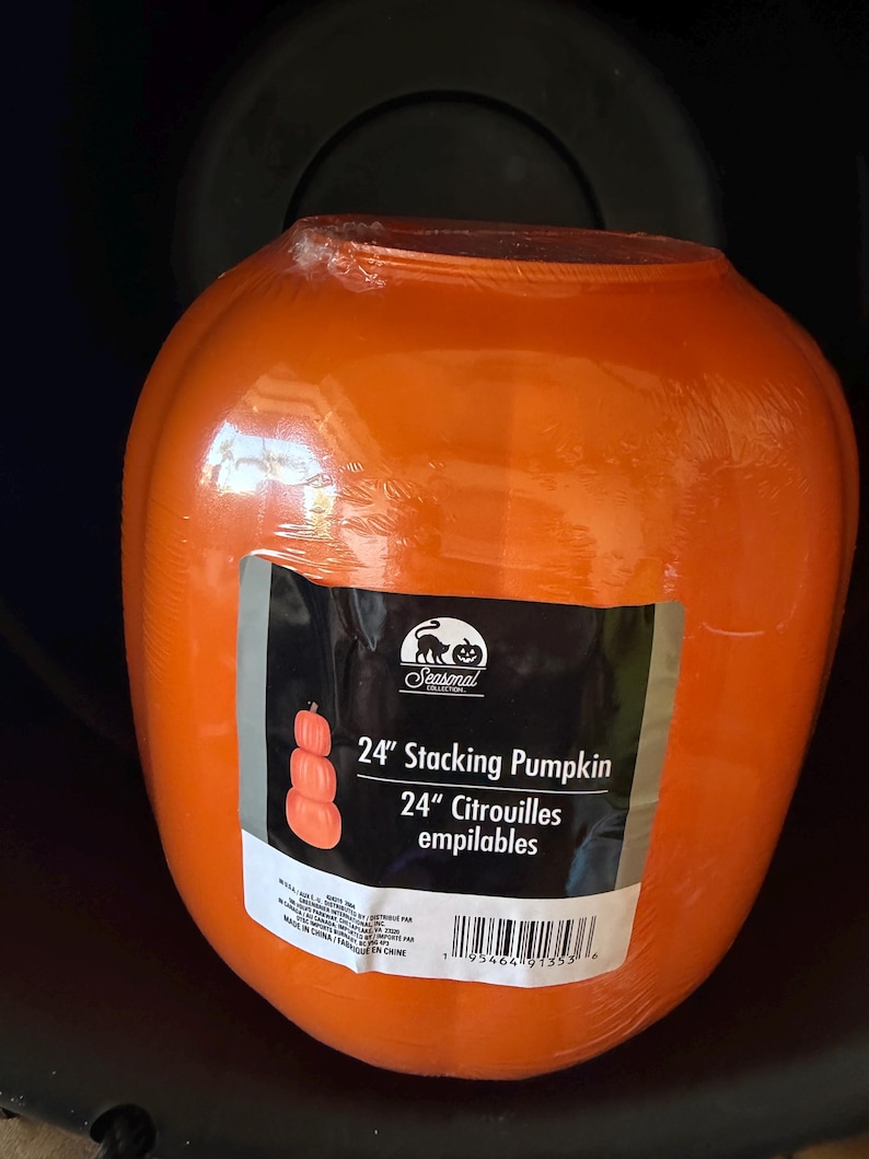 May include: A bright orange, rounded plastic pumpkin decoration wrapped in clear protective film. A rectangular label on the front features a black cat logo, the text 24" Stacking Pumpkin, and a graphic showing three stacked pumpkins.