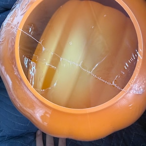 May include: A bright orange plastic pumpkin-shaped container with a smooth, rounded exterior and a wide, circular opening at the top. The opening is covered by a thin, clear, crinkled plastic film that is partially torn across the center.