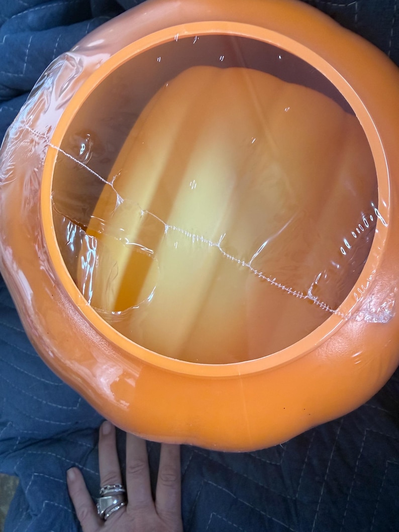 May include: A bright orange plastic pumpkin-shaped container with a smooth, rounded exterior and a wide, circular opening at the top. The opening is covered by a thin, clear, crinkled plastic film that is partially torn across the center.