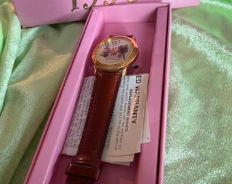 May include: A vintage "Aristocats 1996" wristwatch in a pink box. The watch has a gold-tone case and a brown leather band. The watch face features a scene from the animated film. The box has gold lettering.