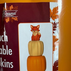 May include: A decorative figurine featuring a small orange and white fox sitting atop a stack of two pumpkins. The top pumpkin is light beige, and the bottom pumpkin is bright orange. The design is displayed on a product label with autumn leaves.