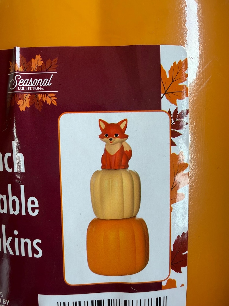 May include: A decorative figurine featuring a small orange and white fox sitting atop a stack of two pumpkins. The top pumpkin is light beige, and the bottom pumpkin is bright orange. The design is displayed on a product label with autumn leaves.