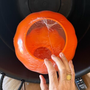 May include: A bright orange, pumpkin-shaped plastic container with a clear plastic film covering the top opening. The pumpkin is nested inside a large, black, round container with black rope handles. A black remote control is visible nearby.