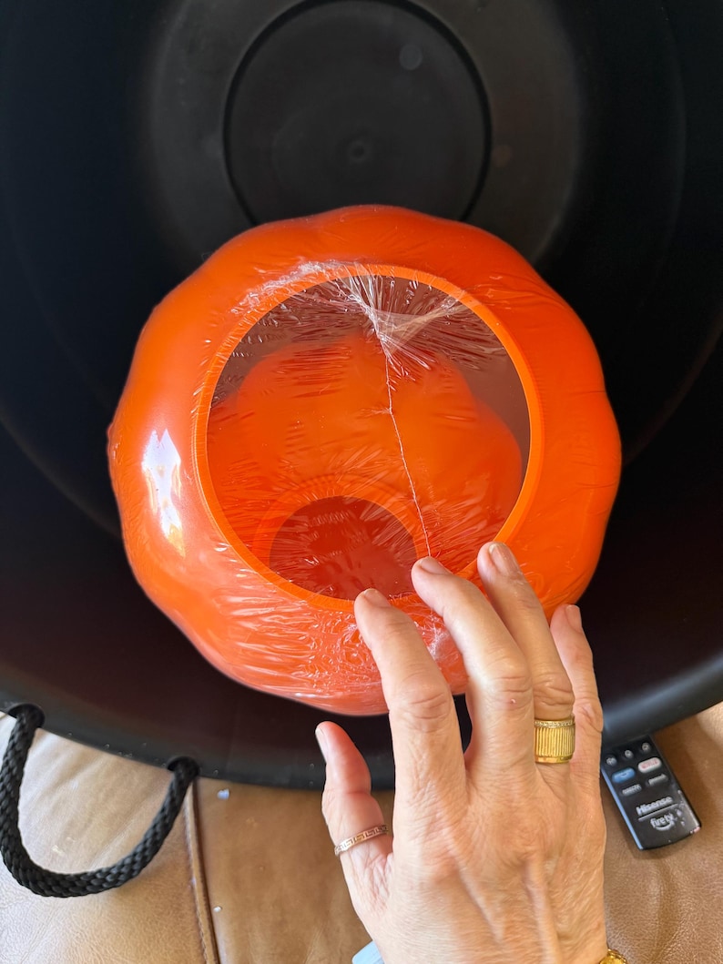 May include: A bright orange, pumpkin-shaped plastic container with a clear plastic film covering the top opening. The pumpkin is nested inside a large, black, round container with black rope handles. A black remote control is visible nearby.
