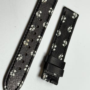 May include: Black leather watch strap with a white floral pattern. The strap has a buckle and is designed for a wristwatch.