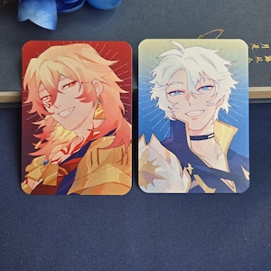 May include: Two rectangular character art cards. One card features a smiling character with golden hair, red eyes, and gold armor. The other card shows a character with white hair, blue eyes, and a blue and gold outfit. Both cards have a colorful background.