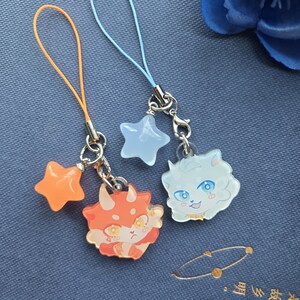 May include: Two charm keychains with cartoon character designs. One keychain has an orange star and a red and orange dinosaur character. The other keychain has a light blue star and a white cat character with blue eyes.