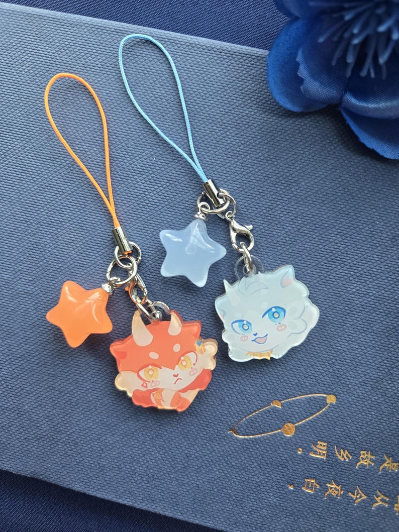 May include: Two charm keychains with cartoon character designs. One keychain has an orange star and a red and orange dinosaur character. The other keychain has a light blue star and a white cat character with blue eyes.