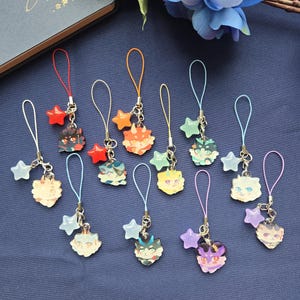 May include: A collection of colorful anime-style charms with various animal faces. Each charm is attached to a silver chain with a star-shaped accent and a loop for attachment. The charms are in shades of red, orange, green, blue, yellow, and purple.