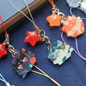 May include: A collection of colorful charms with various designs, including stars and character illustrations. The charms are attached to silver-colored metal chains and colorful cords. The background is a dark blue fabric.