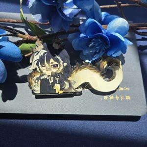 May include: An acrylic pin featuring a stylized anime-inspired character with dark, flowing hair, pointed ears, and a long, pale, curved tail. The character wears a dark outfit with gold accents and has a pensive expression with brown eyes.