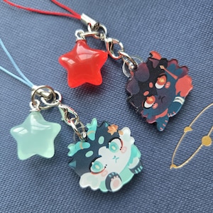 May include: Three decorative charms with silver-colored clasps and cords. One charm is a red star, another is a light green star, and the third is a cartoon character with teal, black, and white accents.