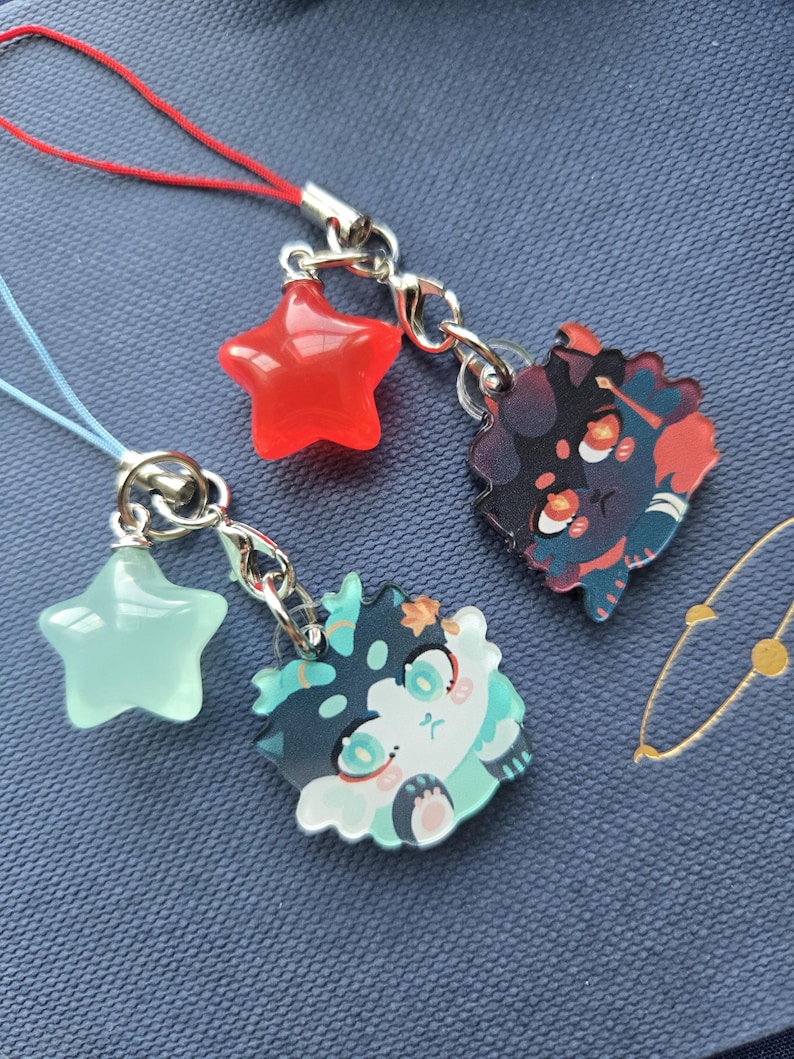 May include: Three decorative charms with silver-colored clasps and cords. One charm is a red star, another is a light green star, and the third is a cartoon character with teal, black, and white accents.