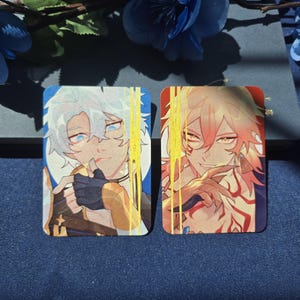 May include: Two rectangular cards featuring anime-style character illustrations. The left card shows a character with white hair and blue eyes, while the right card depicts a character with reddish hair, golden eyes, and red markings. Both have gold foil.