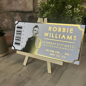 May include: A gold and white ticket stub for a Robbie Williams concert at the Scottish Gas Murrayfield on Saturday, May 31st, 2025. The ticket is for Section E10, Row P, Seat 5/6.