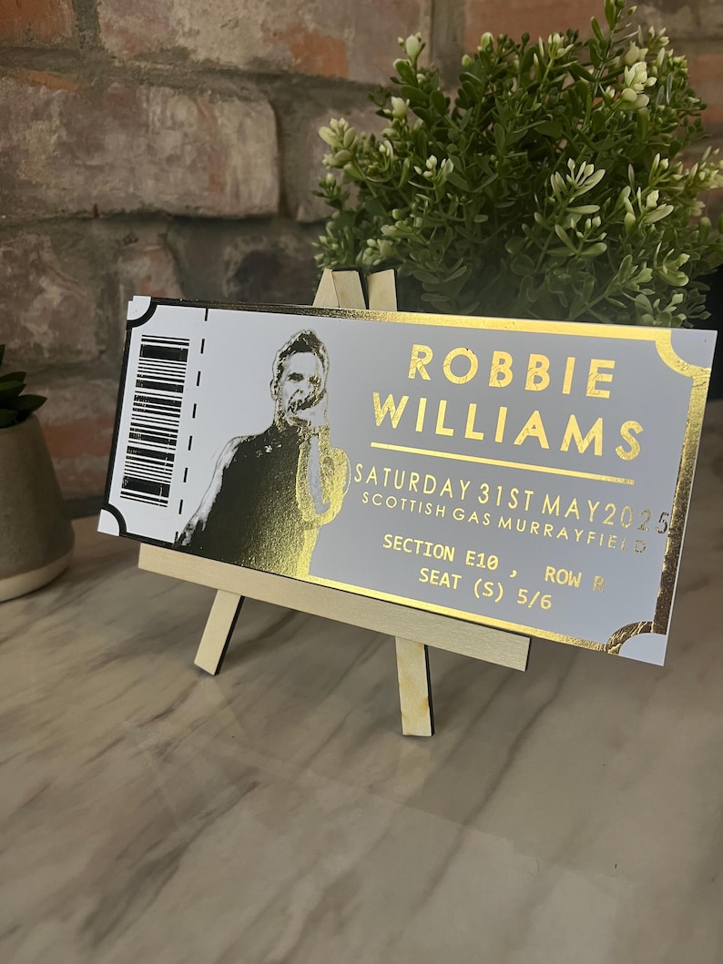 May include: A gold and white ticket stub for a Robbie Williams concert at the Scottish Gas Murrayfield on Saturday, May 31st, 2025. The ticket is for Section E10, Row P, Seat 5/6.