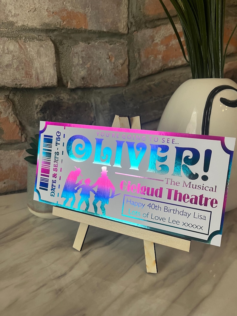 May include: A holographic ticket for the musical "Oliver!" at the Cigud Theatre. The ticket is pink, blue, and purple with a black and white silhouette of characters from the musical. The text on the ticket reads "You're going to see... Oliver! The Musical Cigud Theatre Happy 40th Birthday Lisa Lots of Love Lee xxxxx".
